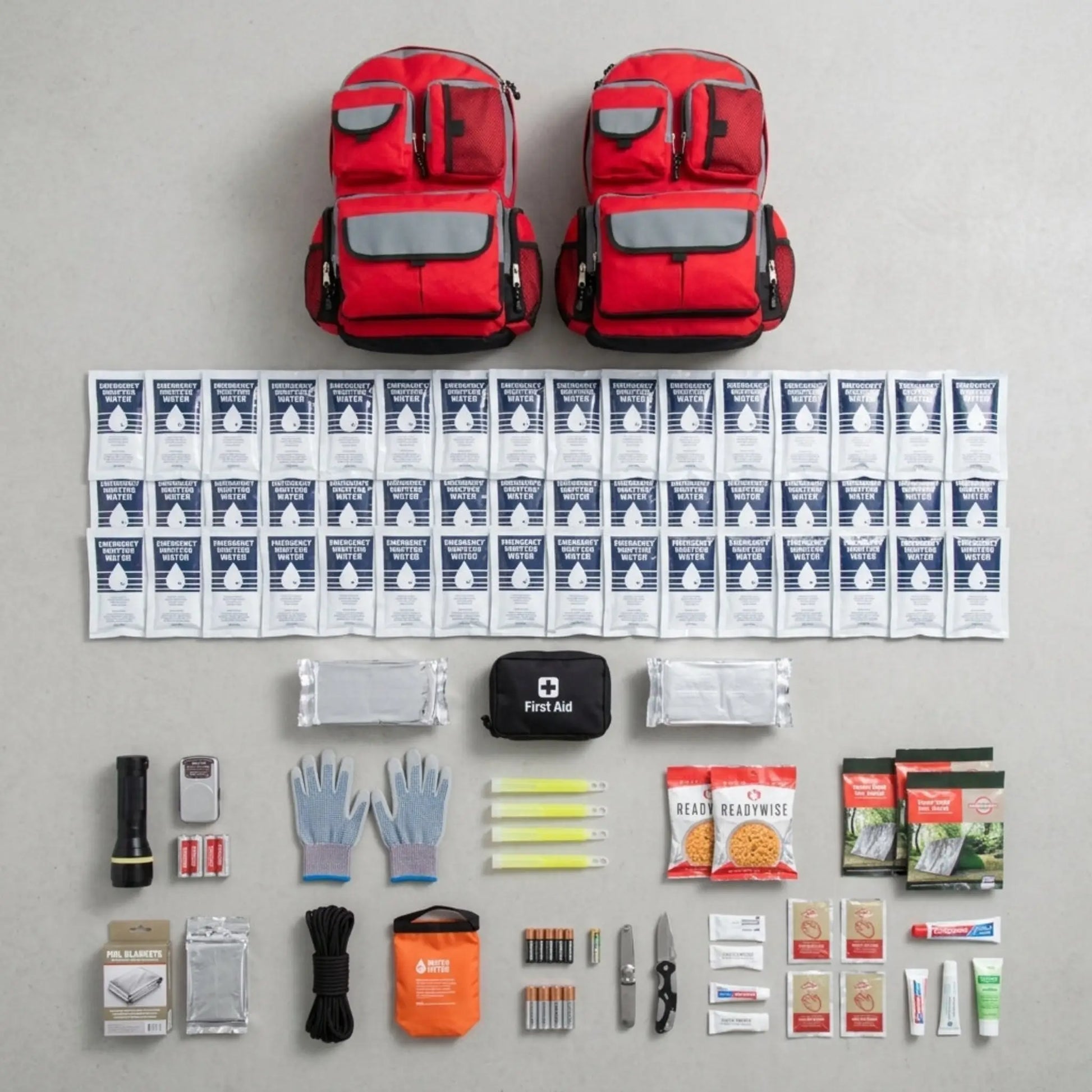 Emergency preparedness kit with red backpacks, first aid supplies, food packets, and other essentials on a gray background.
