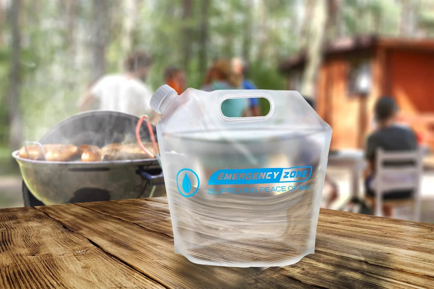 A clear plastic emergency water filter pitcher with a blue and white 'EMERGENCY FILTER' logo, shown on a rustic wooden table with a blurred outdoor campsite background.