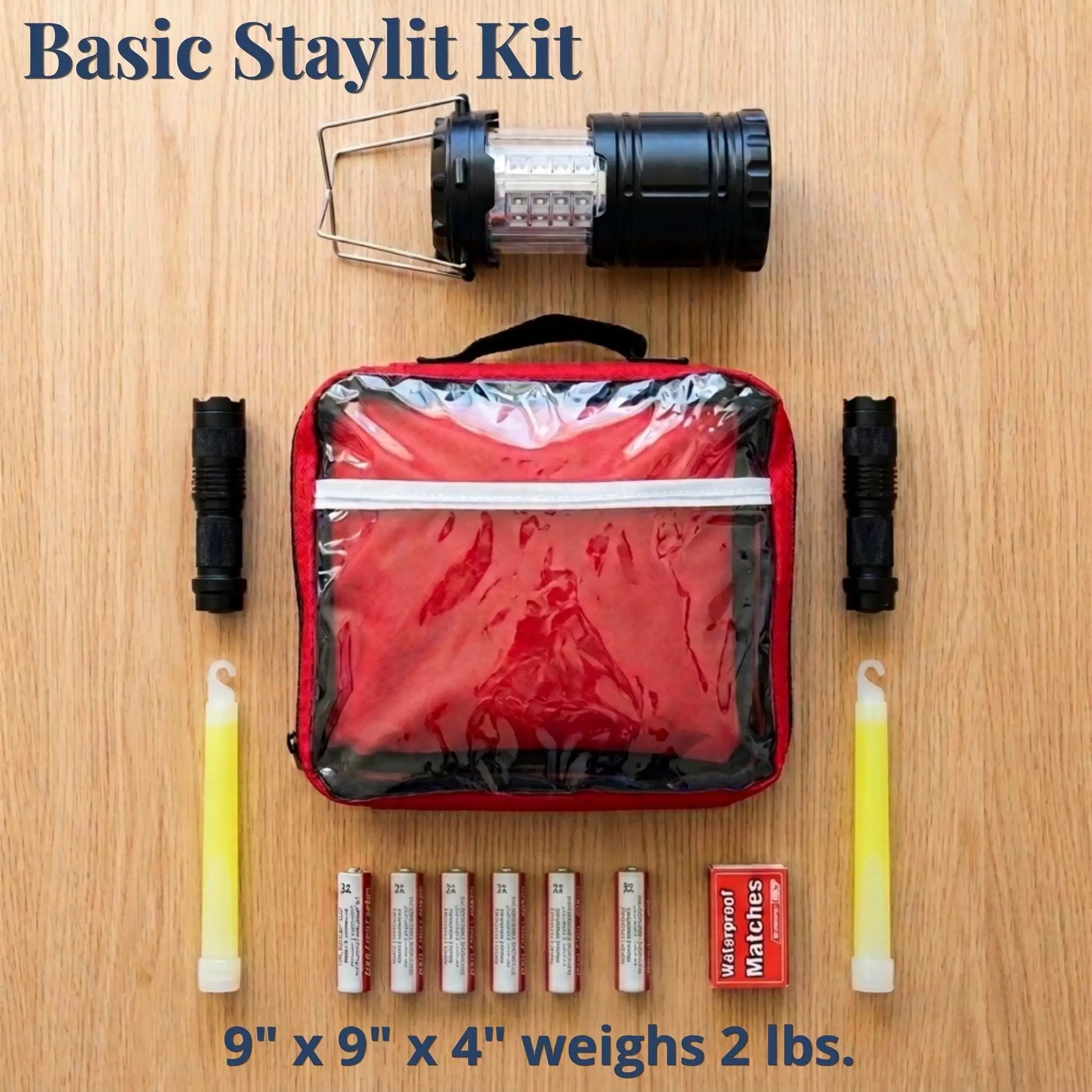 Basic Staylit Kit laid out on a wood surface, including a black lantern, a red zippered carrying case, two black sticks, two yellow glow sticks, six batteries, and a small red waterproof matchbox. Text at the top reads 'Basic Staylit Kit' and text at the bottom says '9" x 9" x 4" weighs 2 lbs.