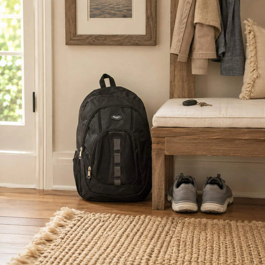 A black backpack with a vertical mesh pocket and a small circular logo on the front, standing on a woven rug next to a wooden bench in a home entryway.